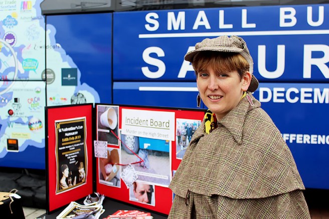 A picture of Jo Smelder, wearing a Sherlock Holmes outfit, in front of the table with her products.